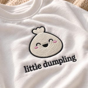 May include: A white long-sleeve sweatshirt featuring an embroidered graphic of a smiling dumpling with pink cheeks. Below the dumpling, the text "little dumpling" is embroidered in black thread. The garment has a ribbed crew neckline.