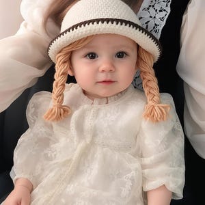 May include: A baby wearing a white crocheted hat with a brown brim and matching yarn braids. The baby is also wearing a white lace dress with puffy sleeves. The baby is looking directly at the camera.