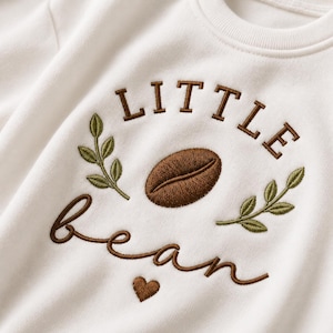 Może przedstawiać: Kremowa bluza z brązowym haftowanym napisem LITTLE bean. Wzór zawiera centralnie umieszczone brązowe ziarno kawy, dwie zielone gałązki z liśćmi po obu stronach oraz małe brązowe serce wyhaftowane pod słowem bean.