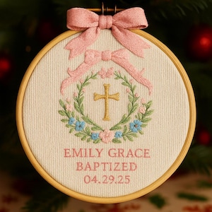 May include: Embroidered baptism ornament in a wooden hoop. The design features a gold cross, a floral wreath with pink and blue flowers, and a pink bow. The text reads "EMILY GRACE BAPTIZED 04.29.25".