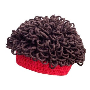 May include: A novelty crochet hat with a brown, curly hair-like design atop a red brim. The hat is handmade and likely intended for costume or playful wear. The brown portion resembles a wig.