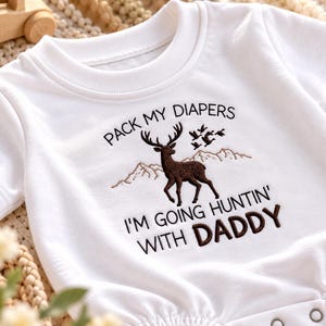May include: A white short-sleeved baby bodysuit featuring an embroidered brown deer, mountain outlines, and flying birds. The text reads: PACK MY DIAPERS I'M GOING HUNTIN' WITH DADDY. The garment has a ribbed crew neckline and metal snap closures.