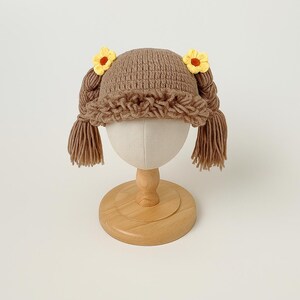 May include: A brown crocheted hat with attached pigtails and yellow flower accents. The hat is displayed on a mannequin head with a wooden stand. The pigtails are made of the same brown yarn as the hat.