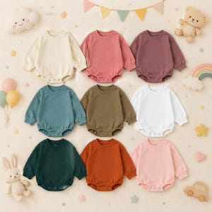 Pode incluir: Uma coleção de nove bodies de bebé de manga comprida em várias cores sólidas, incluindo creme, rosa, roxo escuro, azul-petróleo, verde-azeitona, branco, verde escuro, laranja ferrugem e rosa claro, com gola redonda e botões de pressão.