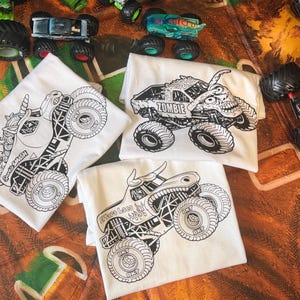 May include: White t-shirts with black monster truck designs. One shirt features a unicorn monster truck with the text "Smash". Another shirt has a zombie monster truck with the text "Zombie". The third shirt has a bull monster truck with the text "Where Lace".