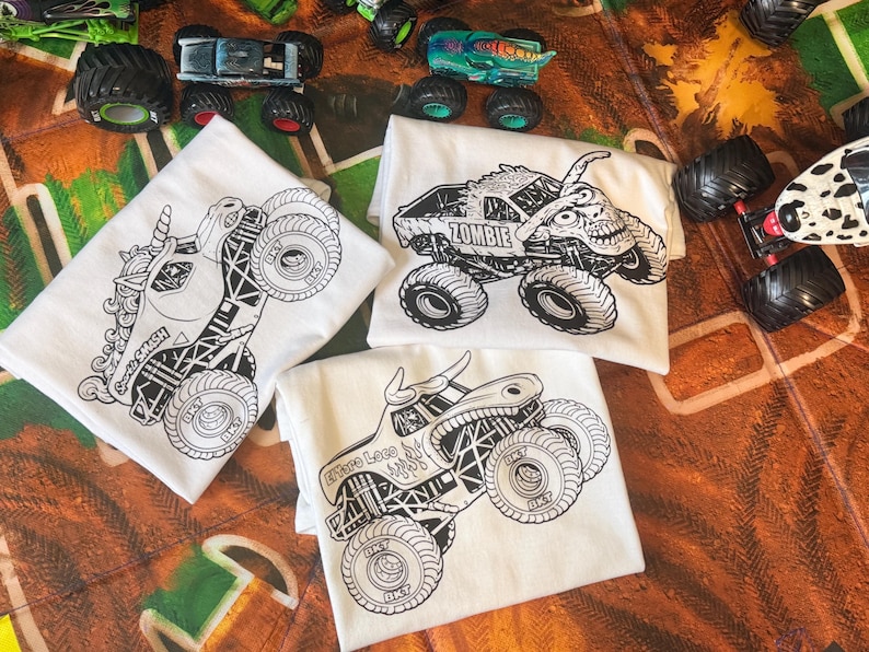 May include: White t-shirts with black monster truck designs. One shirt features a unicorn monster truck with the text "Smash". Another shirt has a zombie monster truck with the text "Zombie". The third shirt has a bull monster truck with the text "Where Lace".