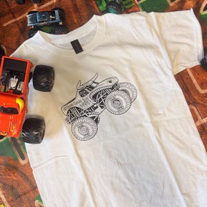 May include: White t-shirt featuring a black and white monster truck graphic with the text "Grave Loco". Several toy monster trucks in various colors surround the shirt, suggesting a theme of monster truck toys and apparel.