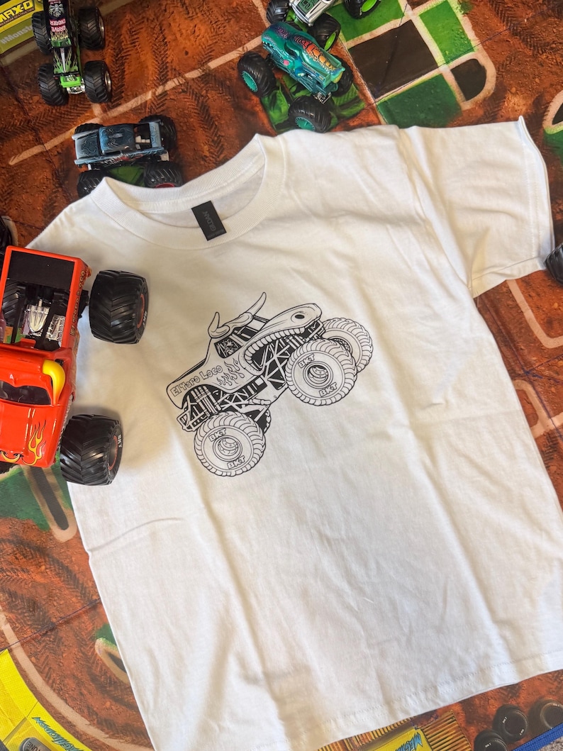 May include: White t-shirt featuring a black and white monster truck graphic with the text "Grave Loco". Several toy monster trucks in various colors surround the shirt, suggesting a theme of monster truck toys and apparel.