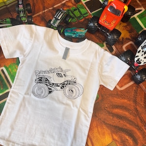 May include: White t-shirt featuring a black and white graphic of a monster truck with a unicorn horn and the text "Sweet Smash". Several toy monster trucks in various colors surround the shirt.