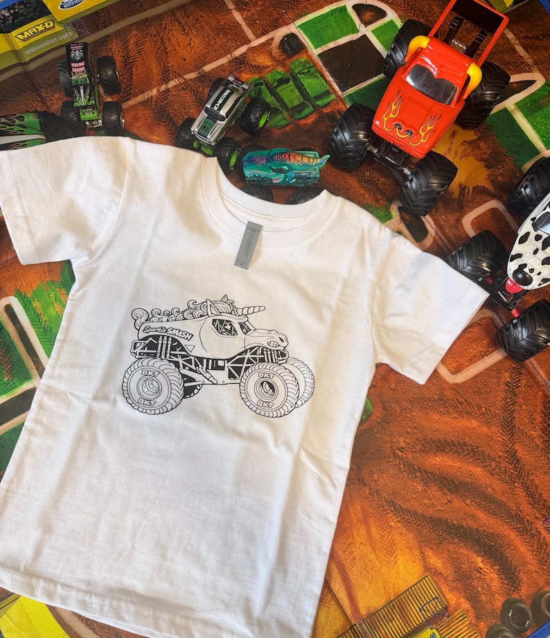May include: White t-shirt featuring a black and white graphic of a monster truck with a unicorn horn and the text "Sweet Smash". Several toy monster trucks in various colors surround the shirt.