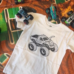 May include: White t-shirt featuring a black and white graphic of a monster truck with the word "ZOMBIE" on it. Several toy monster trucks in various colors are scattered around the shirt on a brown and green play mat.