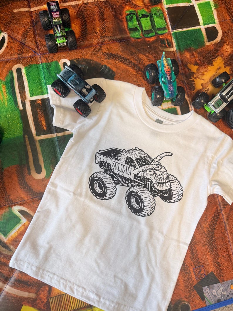 May include: White t-shirt featuring a black and white graphic of a monster truck with the word "ZOMBIE" on it. Several toy monster trucks in various colors are scattered around the shirt on a brown and green play mat.