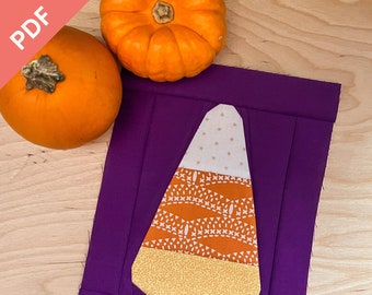Puede incluir: Un cuadrado de tela morado con una aplicación de maíz dulce hecha de diferentes telas estampadas. El maíz dulce es blanco, naranja y amarillo. Hay dos calabazas naranjas en el fondo.