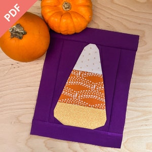 May include: A purple fabric square with a candy corn applique made from different patterned fabrics. The candy corn is white, orange, and yellow. Two orange pumpkins are in the background.