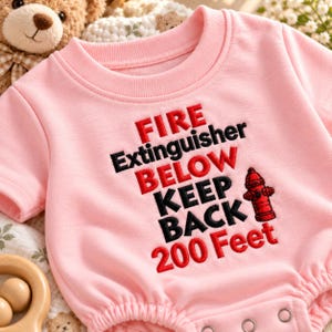 May include: A light pink short-sleeved bodysuit featuring embroidered text that reads FIRE Extinguisher BELOW KEEP BACK 200 Feet in red and black thread, accompanied by a small red embroidered fire hydrant icon on the right side.