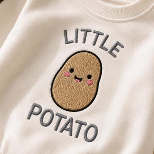 May include: A cream-colored sweatshirt featuring an embroidered graphic of a tan potato with a smiling face and pink cheeks. The text "LITTLE" is embroidered in dark gray above the potato, and the word "POTATO" is embroidered in dark gray below.
