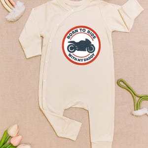 Puede incluir: Mono de bebé color crema con mangas largas. La parte delantera presenta un gráfico circular con una motocicleta y el texto "BORN TO RIDE WITH MY DADDY" en rojo y azul. El mono tiene un cierre envolvente con botones blancos.