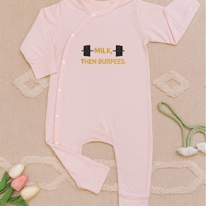 May include: A light pink baby onesie with long sleeves and a wrap-style closure. The onesie features the text "MILK, THEN BURPEES" with a barbell graphic in black. The garment is made of soft material and is designed for comfort.