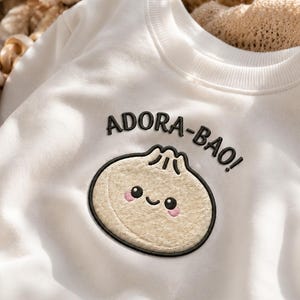 Puede incluir: Una sudadera blanca de manga larga con un parche bordado de una bola de masa beige con cara sonriente y mejillas rosadas. Sobre la bola de masa, el texto ADORA-BAO! está bordado en hilo negro. La prenda tiene un cuello redondo acanalado.