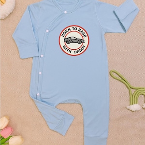 Pode incluir: Macacão de bebê azul claro com mangas compridas. O macacão apresenta um emblema redondo com o texto "BORN TO RACE WITH DADDY" e um gráfico de carro. A peça tem fecho de botão e é feita de um material macio.