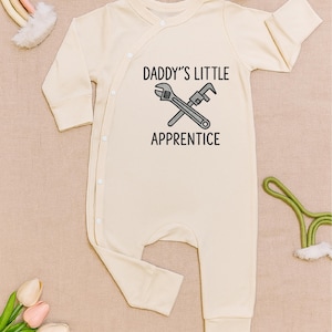 May include: Cream-colored baby onesie with long sleeves and a wrap-style closure. The front features the text "DADDY'S LITTLE APPRENTICE" above a crossed wrench and pipe wrench graphic in gray.
