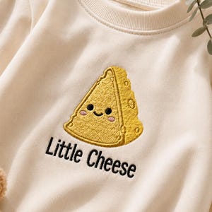 May include: A cream-coloured crewneck sweatshirt featuring an embroidered yellow wedge of cheese with a smiling face and pink cheeks. Below the cheese, the text Little Cheese is embroidered in black thread, centred on the front of the garment.