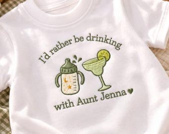 Könnte beinhalten: Ein weißes kurzärmeliges T-Shirt mit grüner Stickerei eines Babyfläschchens und eines Margaritaglases. Der Text lautet I'd rather be drinking with Aunt Jenna, ergänzt durch ein kleines grünes Herzsymbol sowie Mond- und Sternmotive auf der Flasche.