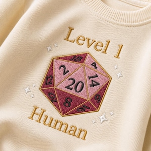 May include: A cream-coloured sweatshirt featuring an embroidered twenty-sided die in shades of pink and burgundy. The text "Level 1" appears above the die and "Human" below it in gold thread. Small white embroidered stars surround the central design.