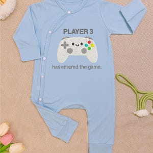 Op de afbeelding: Lichtblauwe baby-onesie met lange mouwen. Het heeft een grijze controller-afbeelding en de tekst "PLAYER 3 has entered the game." De onesie heeft een knoopsluiting en is gemaakt van zacht materiaal.