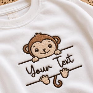 May include: A white long-sleeve sweatshirt featuring an embroidered brown monkey design. The monkey is positioned above and below a horizontal line with the text Your Text embroidered in dark brown thread between the monkey's head and tail.