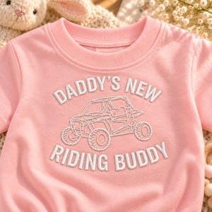 Könnte beinhalten: Ein hellrosa T-Shirt mit kurzen Ärmeln und weißem Sticktext, der DADDY'S NEW RIDING BUDDY lautet. Zentriert zwischen dem Text befindet sich die weiße Stickkontur eines Geländefahrzeugs mit großen Reifen und einem Überrollkäfig-Rahmen.