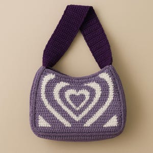 May include: A crocheted handbag in shades of purple and white. The bag features a heart design on the front and a solid purple strap. The bag is a small, shoulder-style bag.
