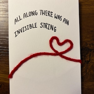 Könnte beinhalten: Eine weiße Grußkarte mit dem schwarzen Text "ALL ALONG THERE WAS AN INVISIBLE STRING". Ein roter Garnfaden formt ein Herz und verläuft über die Karte, was dem Design eine taktile Note verleiht. Schlichtes, gefühlvolles Design.