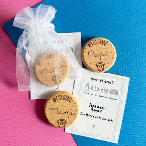 Può includere: Gettoni in legno incisi con le parole "Mama" e "Papa", accompagnati da un piccolo sacchetto bianco e una carta con testo tedesco e icone per bambini. Lo sfondo è rosa e blu, creando una presentazione colorata.