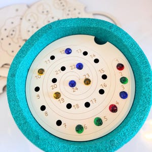 Può includere: Un giocattolo in legno turchese con un design a spirale e gemme colorate. Il giocattolo ha numeri attorno alla spirale, con punti neri e gemme colorate. Ulteriori pezzi di legno sono visibili sullo sfondo.