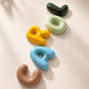 Puede incluir: Un conjunto de cinco formas cerámicas tridimensionales, lisas y redondeadas, en varios colores que incluyen verde oscuro, verde claro, amarillo, azul claro y marrón. Cada pieza tiene un acabado brillante y una forma orgánica, suave y abultada.
