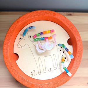 Pode incluir: Uma bandeja de plástico laranja com uma inserção de madeira com um desenho a traço de um cavalo. Limpadores de cachimbo coloridos com contas são dispostos no cavalo, criando um brinquedo lúdico e educativo. A bandeja tem duas alças recortadas.