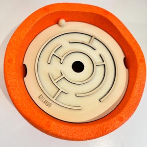 May include: An orange and cream-colored wooden maze puzzle with a small white ball. The maze is circular, with a series of pathways and a central hole. The outer rim is orange, and the inner maze is cream-colored. The name "RICCARDO" is printed on the maze.
