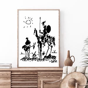Op de afbeelding: Zwart-wit kunstafdruk met een ridder te paard met een speer, een metgezel en windmolens, onder een zon. Het kunstwerk is in minimalistische stijl, ingelijst in een licht houten frame en weergegeven op een houten kast.