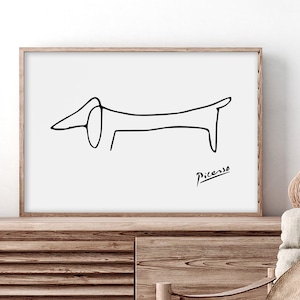 May include: Black and white line drawing of a dachshund, signed "Picasso" in the bottom right corner. The drawing is framed and displayed on a wooden shelf.