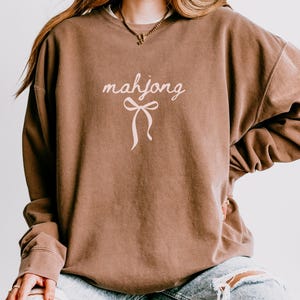 May include: A brown sweatshirt with the word "mahjong" in a cursive font and a bow graphic. The sweatshirt has long sleeves and a relaxed fit. The garment is a solid brown color.