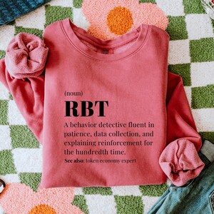 May include: A dusty rose sweatshirt with the letters "RBT" in black, with text below defining the term. The sweatshirt is folded with a matching scrunchie. The background includes a green and white checkered pattern and a pink flower.