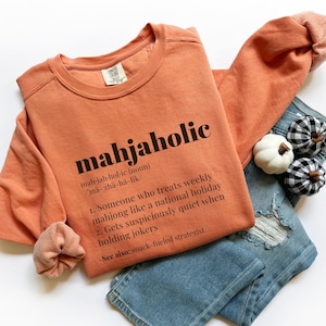 May include: An orange sweatshirt with the word "mahjaholic" printed in black, along with a definition. The sweatshirt is paired with distressed blue jeans and decorative pumpkins. A scrunchie is also visible.