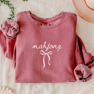 May include: A dusty rose-colored sweatshirt with the word "mahjong" in a cursive font and a bow graphic. The sweatshirt has a crew neck and long sleeves with ribbed cuffs. A matching scrunchie is next to the sweatshirt.