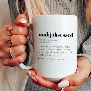May include: White ceramic mug with the word "mahjobsessed" printed in black. The mug features a definition of the word, describing someone who loves mahjong. The mug is held by a person with multiple rings on their fingers.