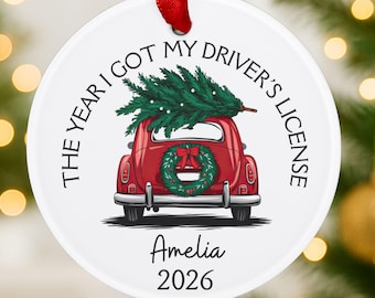 Pode incluir: Um ornamento redondo de cerâmica branca com uma fita vermelha. O design apresenta um carro vermelho transportando um pinheiro verde no teto, uma guirlanda no porta-malas e o texto THE YEAR I GOT MY DRIVER'S LICENSE, Amelia e 2026 em preto.