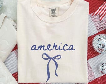 May include: Cream-colored t-shirt with the word "america" in blue script and a blue bow graphic. The shirt is folded on a red and white checkered surface, with white boots and disco balls in the background.