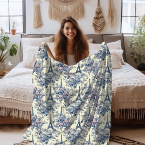 May include: A cream-colored throw blanket with a repeating pattern of the Eiffel Tower and blue and pink floral designs.
