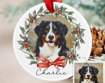 May include: A white ceramic Christmas ornament featuring a Bernese Mountain Dog portrait, surrounded by a green and red holly wreath and a red bow. The name "Charlie" is written in cursive. A red ribbon is attached for hanging.
