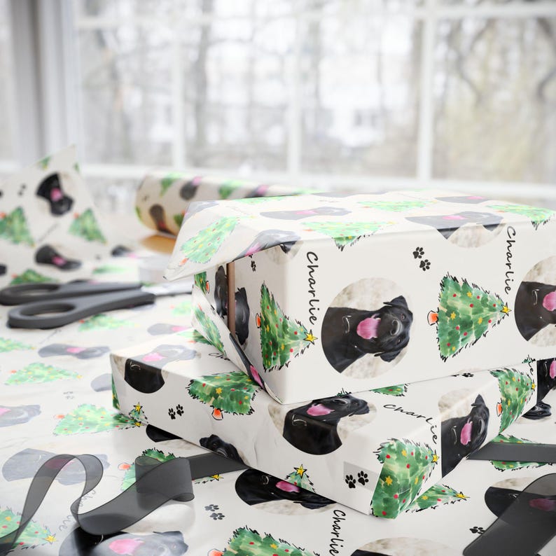 Pode incluir: Papel de embrulho branco com um padr&atilde;o repetido de um c&atilde;o preto, &aacute;rvores de Natal e pegadas. O c&atilde;o est&aacute; num c&iacute;rculo com o nome "Charlie" escrito na lateral. O papel est&aacute; embrulhado em volta de uma caixa de presente. Tesouras e fita tamb&eacute;m s&atilde;o vis&iacute;veis.